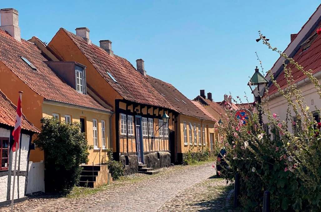 En brosteinsgate omgitt av historiske, gule hus med røde tak og blomstrende planter på siden, under en klar blå himmel.