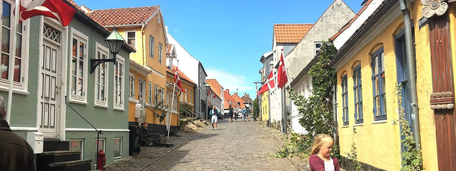 En brosteinsgate omgitt av fargerike hus med danske flagg, hvor folk går og en jente står med en barnevogn under en klar blå himmel.