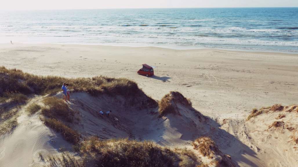 Rød bil står parkert på stranden mens familien hygger seg i sanden.