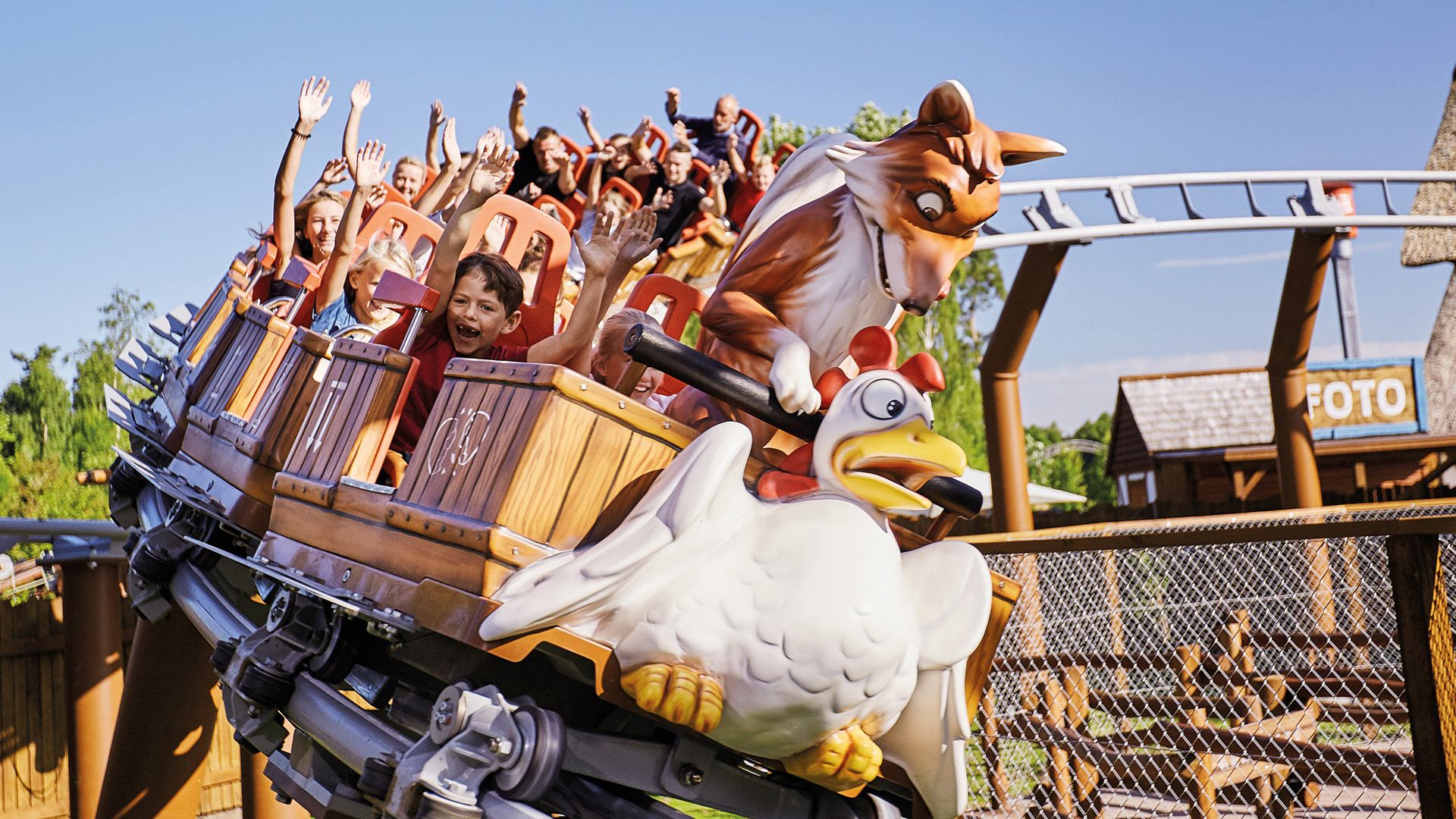 En berg-og-dal-bane med vogner dekorert som tønnene med figurer av en hund og en høne, fylt med glade mennesker som løfter hendene i luften, under en klar blå himmel.