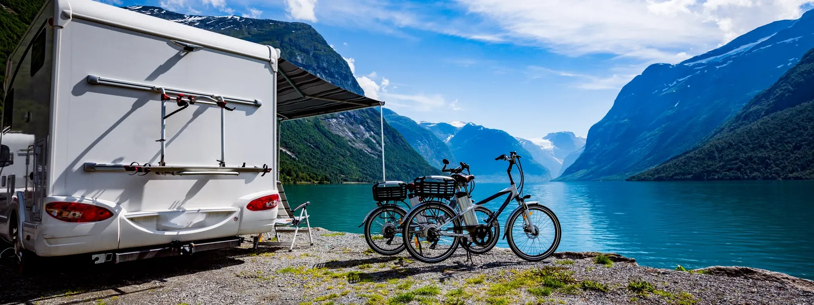 A motorhome and two bicycles parked by a fjord, surrounded by majestic mountains under a bright blue sky.