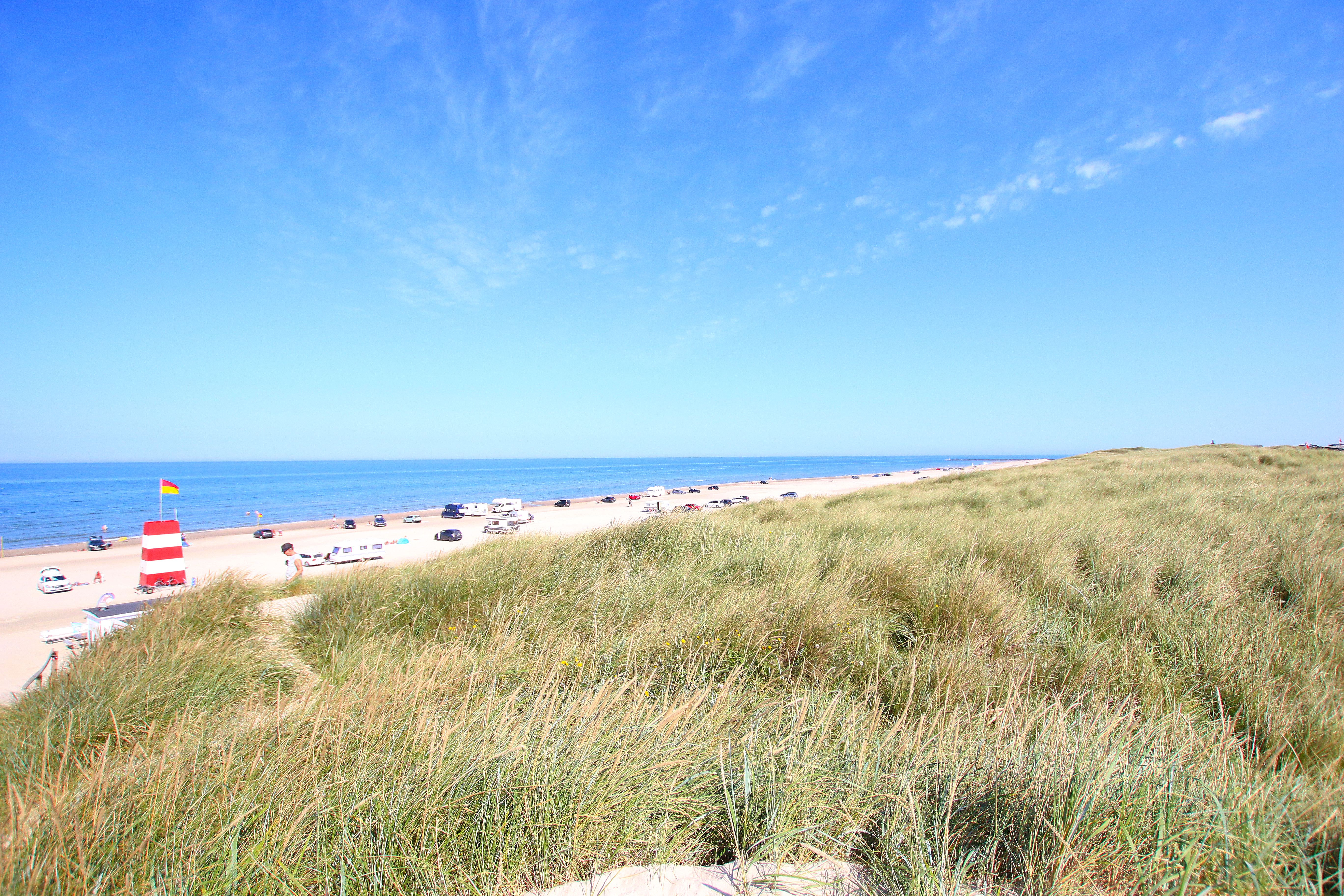 En solrik dag på en sandstrand med gresskledde sanddyner og biler parkert langs stranden. En rød- og hvitstripet livvakttårn flagrer med en gul-rød sikkerhetsflagg.