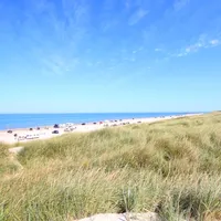En solrik dag på en sandstrand med gresskledde sanddyner og biler parkert langs stranden. En rød- og hvitstripet livvakttårn flagrer med en gul-rød sikkerhetsflagg.