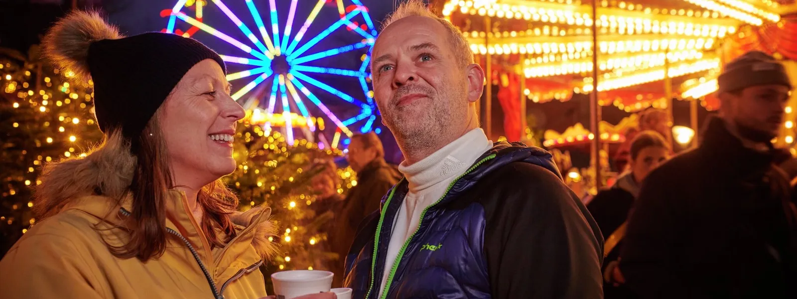 Par (kvinne og mann) som nyter en kopp med noe varmt og drikke foran opplyste karuseller. Foto.