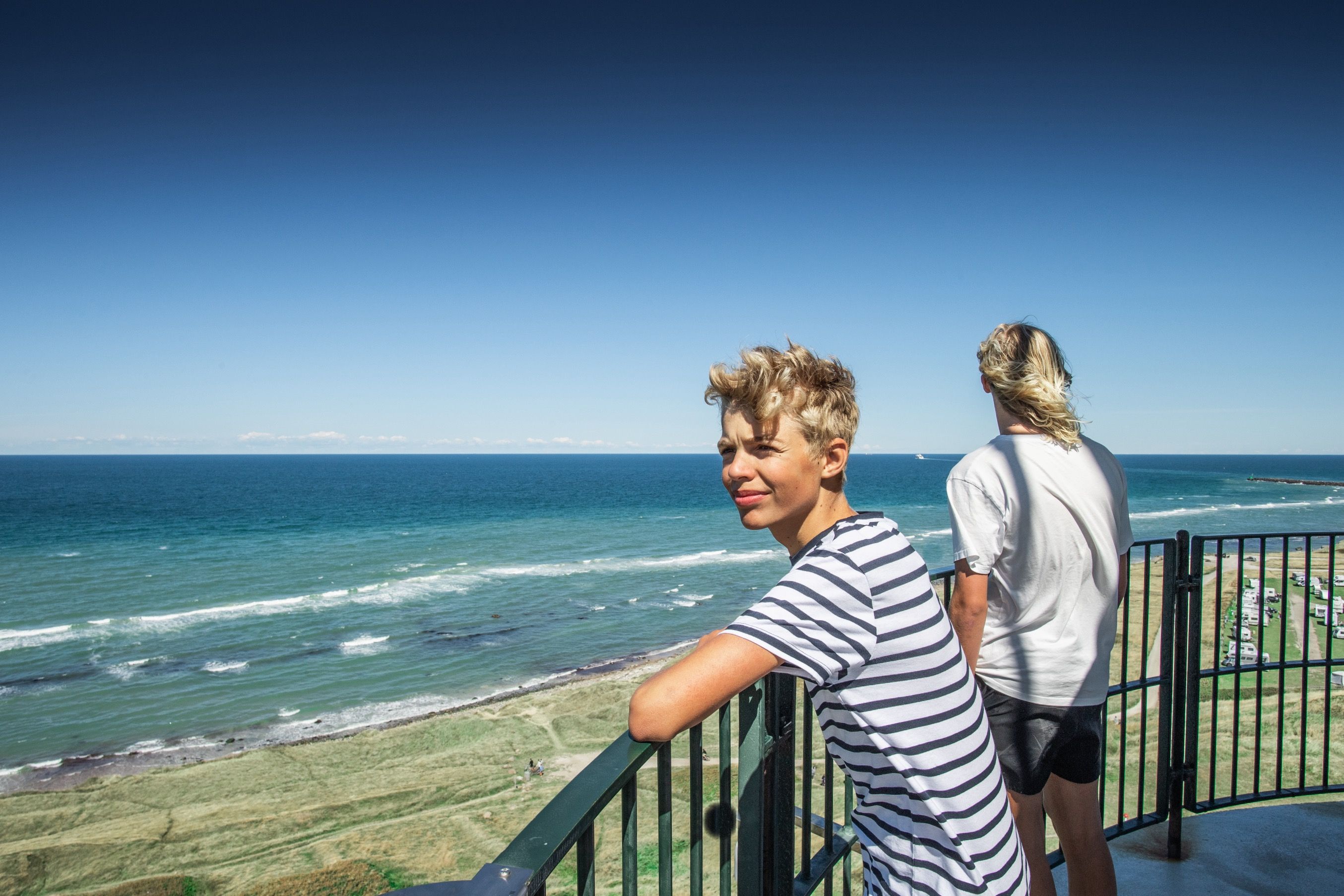 To gutter står på en utkikksplattform og ser ut over havet og stranden under en klar blå himmel.