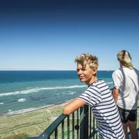 To gutter står på en utkikksplattform og ser ut over havet og stranden under en klar blå himmel.
