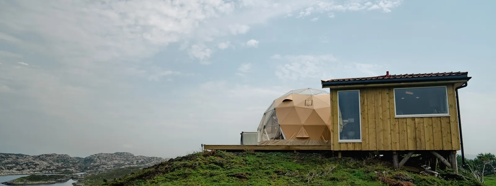 Et moderne kuppeltelt og en lille træhytte beliggende på en grøn bakke med udsigt over klippefyldte landskaber under en let overskyet himmel.