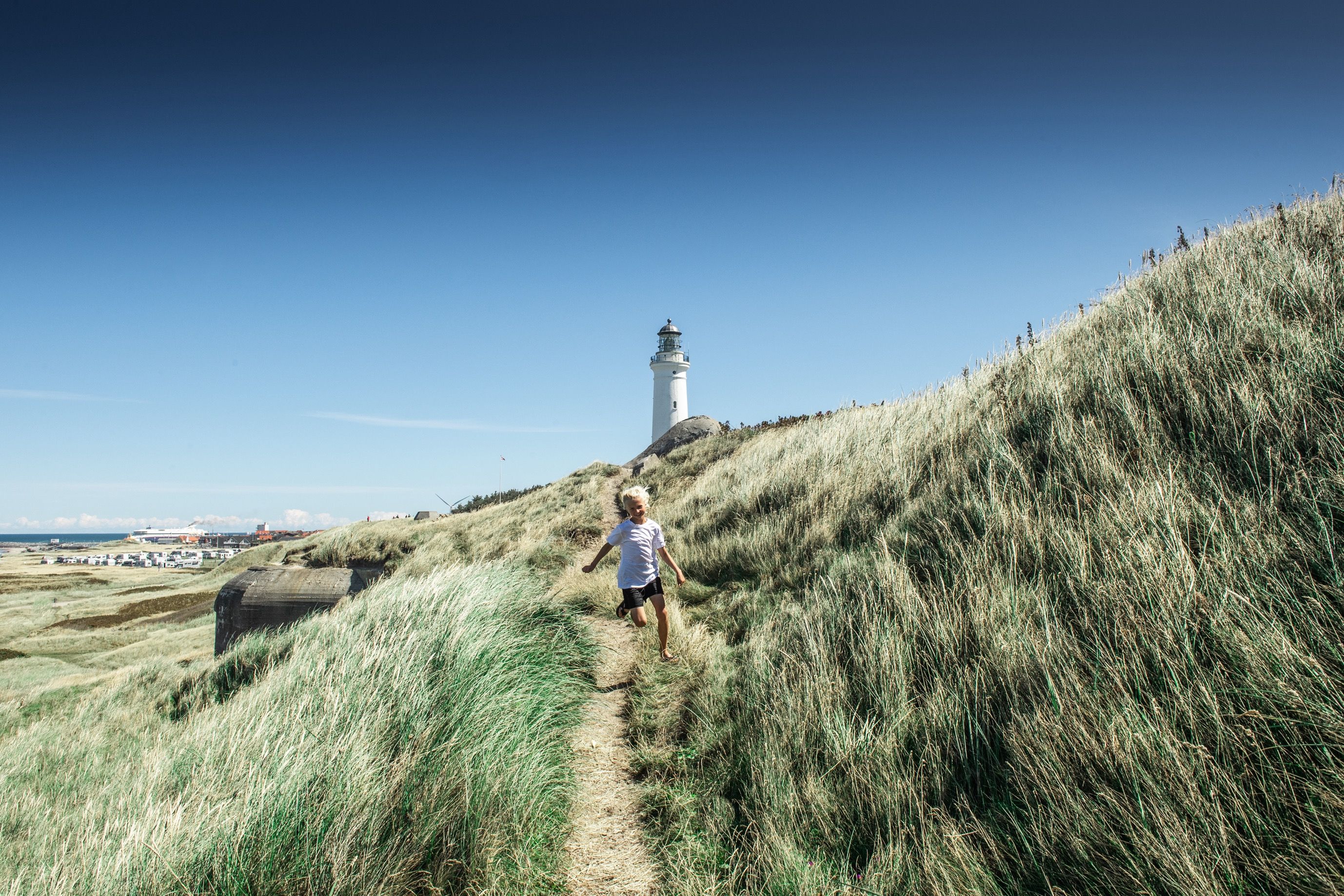 En gutt løper langs en sti gjennom høyt gress med et fyrtårn i bakgrunnen, under en klar blå himmel.