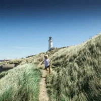 En gutt løper langs en sti gjennom høyt gress med et fyrtårn i bakgrunnen, under en klar blå himmel.