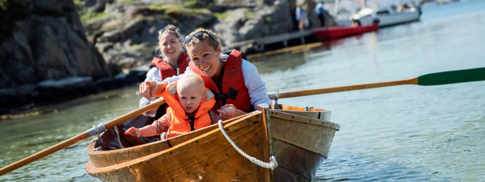 Smiling adults and a small child in life jackets enjoying a rowboat trip along a scenic coastal area with rocky shores and calm water.