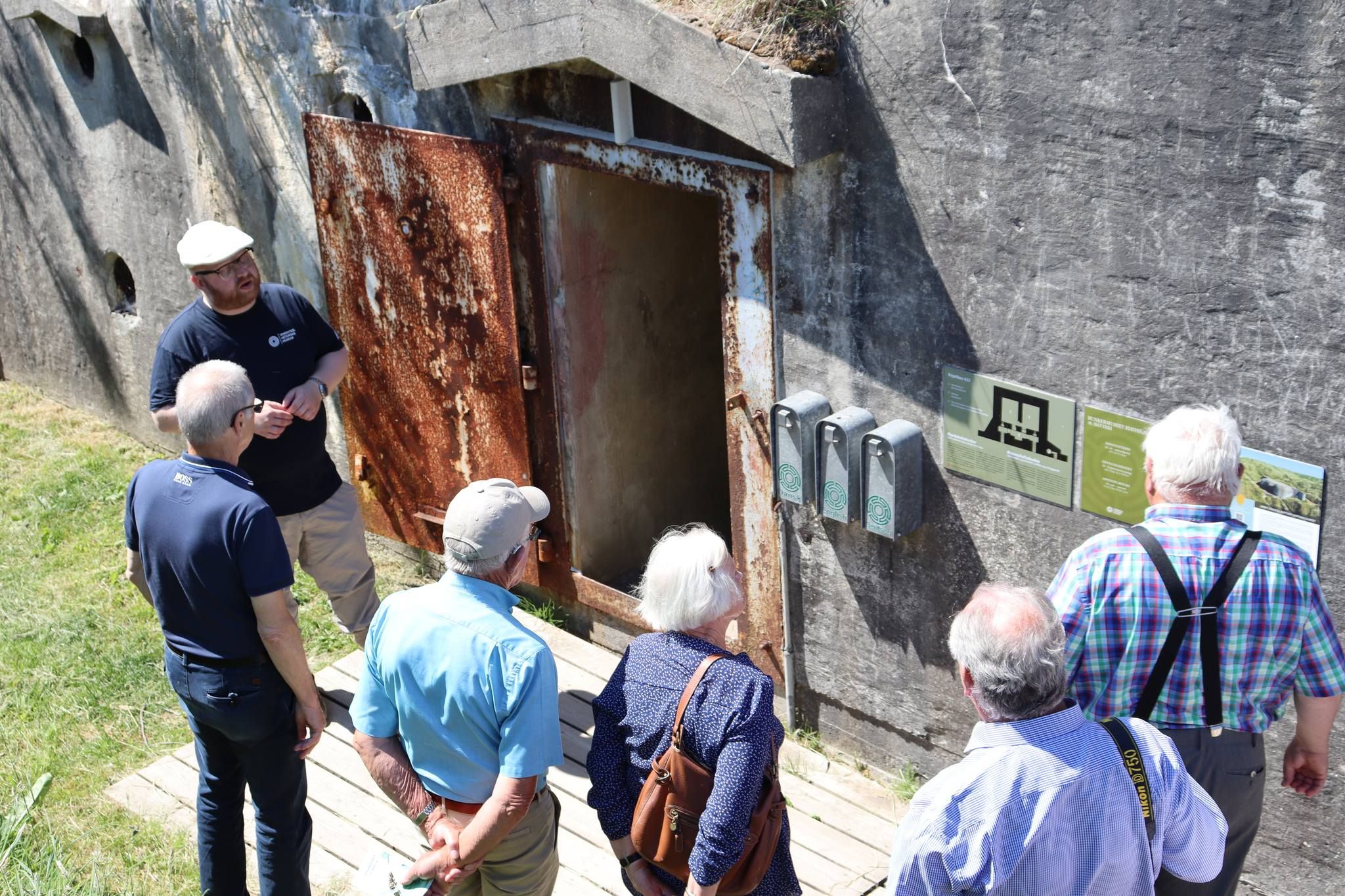 En guide viser en gruppe besøkende en inngang til en gammel bunker. Den rustne døren står åpen, og gruppen lytter oppmerksomt mens guiden forklarer. Flere informasjonsplakater og dispensere er montert på bunkerens betongvegg.