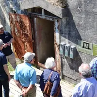 En guide viser en gruppe besøkende en inngang til en gammel bunker. Den rustne døren står åpen, og gruppen lytter oppmerksomt mens guiden forklarer. Flere informasjonsplakater og dispensere er montert på bunkerens betongvegg.