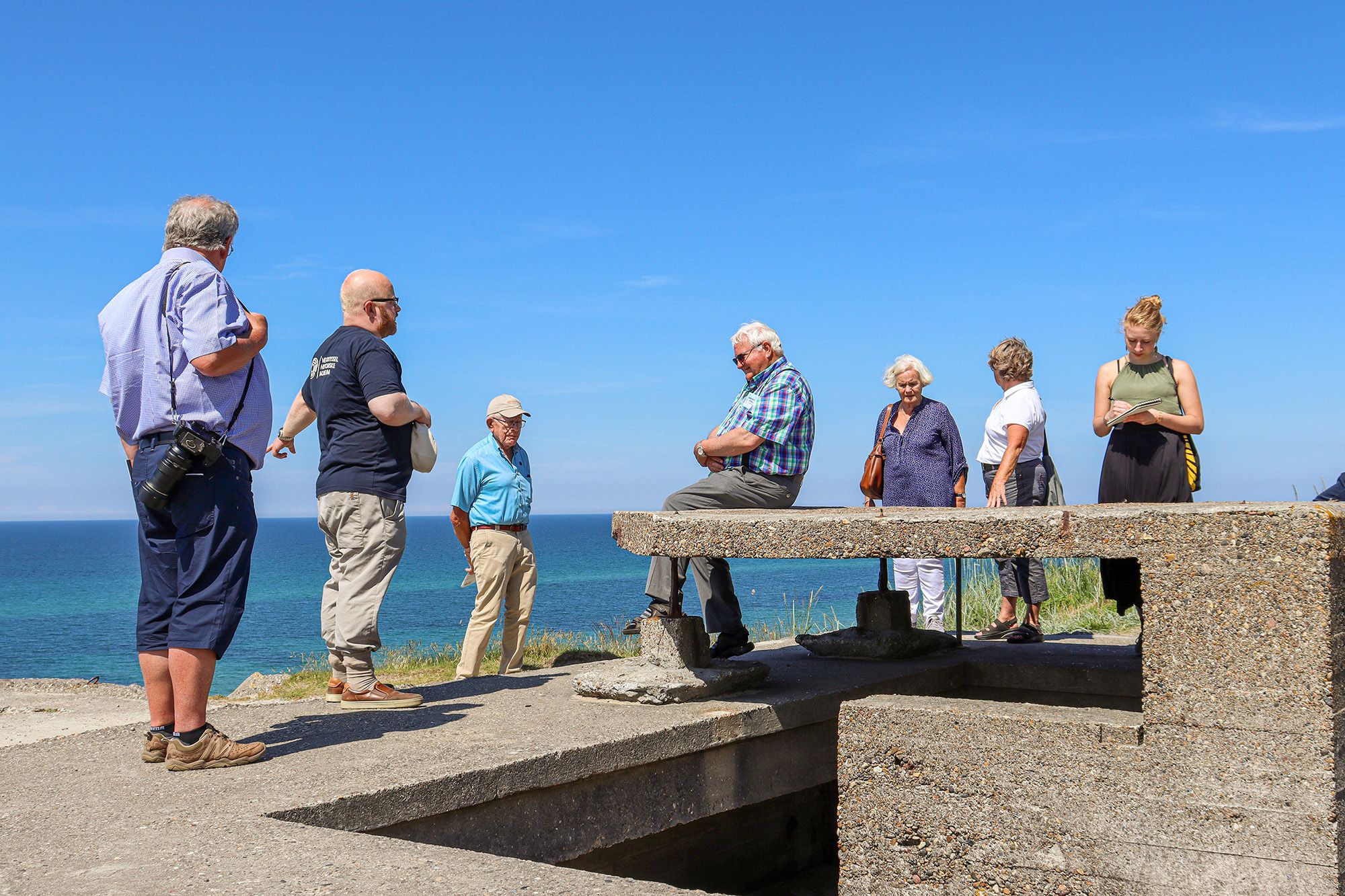 En gruppe mennesker på en guidet tur ved en bunker fra andre verdenskrig. De står og sitter på en betongstruktur med utsikt over havet under en klar blå himmel. En av deltakerne har et kamera rundt halsen, og en annen noterer i en bok.