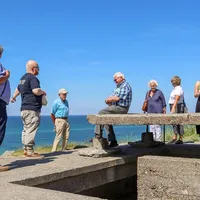 En gruppe mennesker på en guidet tur ved en bunker fra andre verdenskrig. De står og sitter på en betongstruktur med utsikt over havet under en klar blå himmel. En av deltakerne har et kamera rundt halsen, og en annen noterer i en bok.