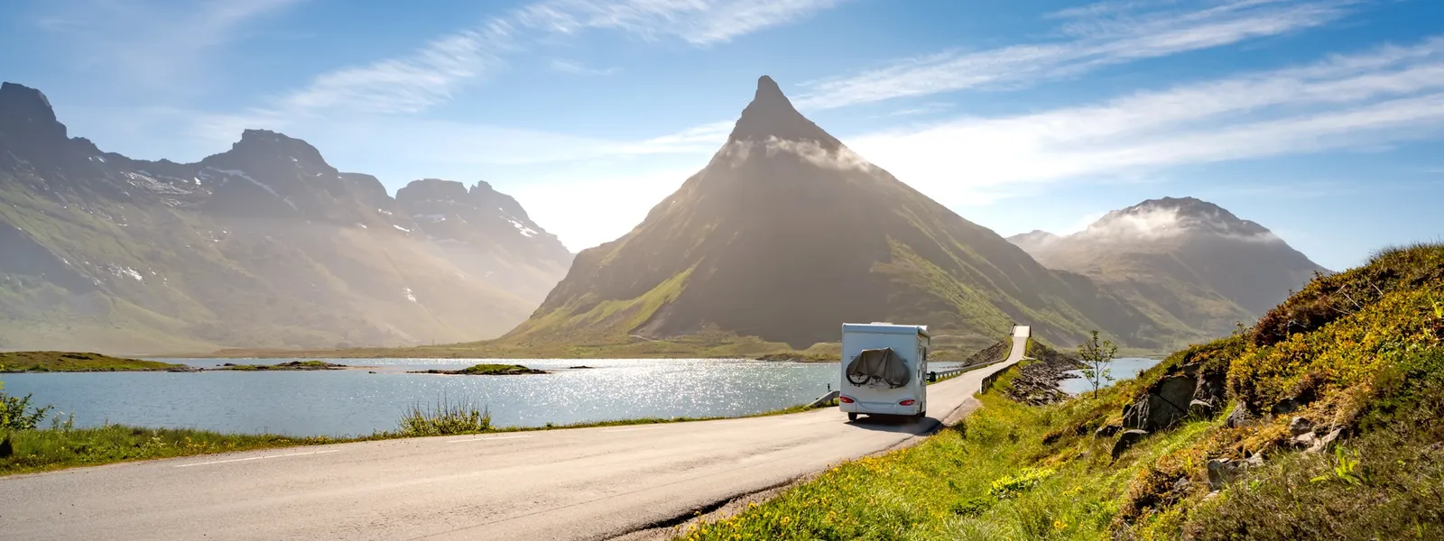 Ein Wohnmobil fährt auf einer Straße entlang eines malerischen Sees, flankiert von beeindruckenden Bergen unter einem strahlend blauen Himmel.