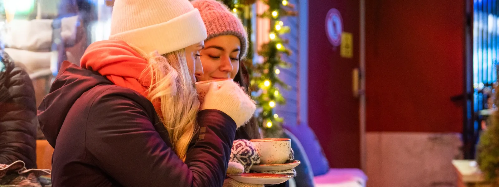 Zwei Frauen genießen ein warmes Getränk, während sie in Winterkleidung eingepackt draußen an einem kalten Abend sitzen.