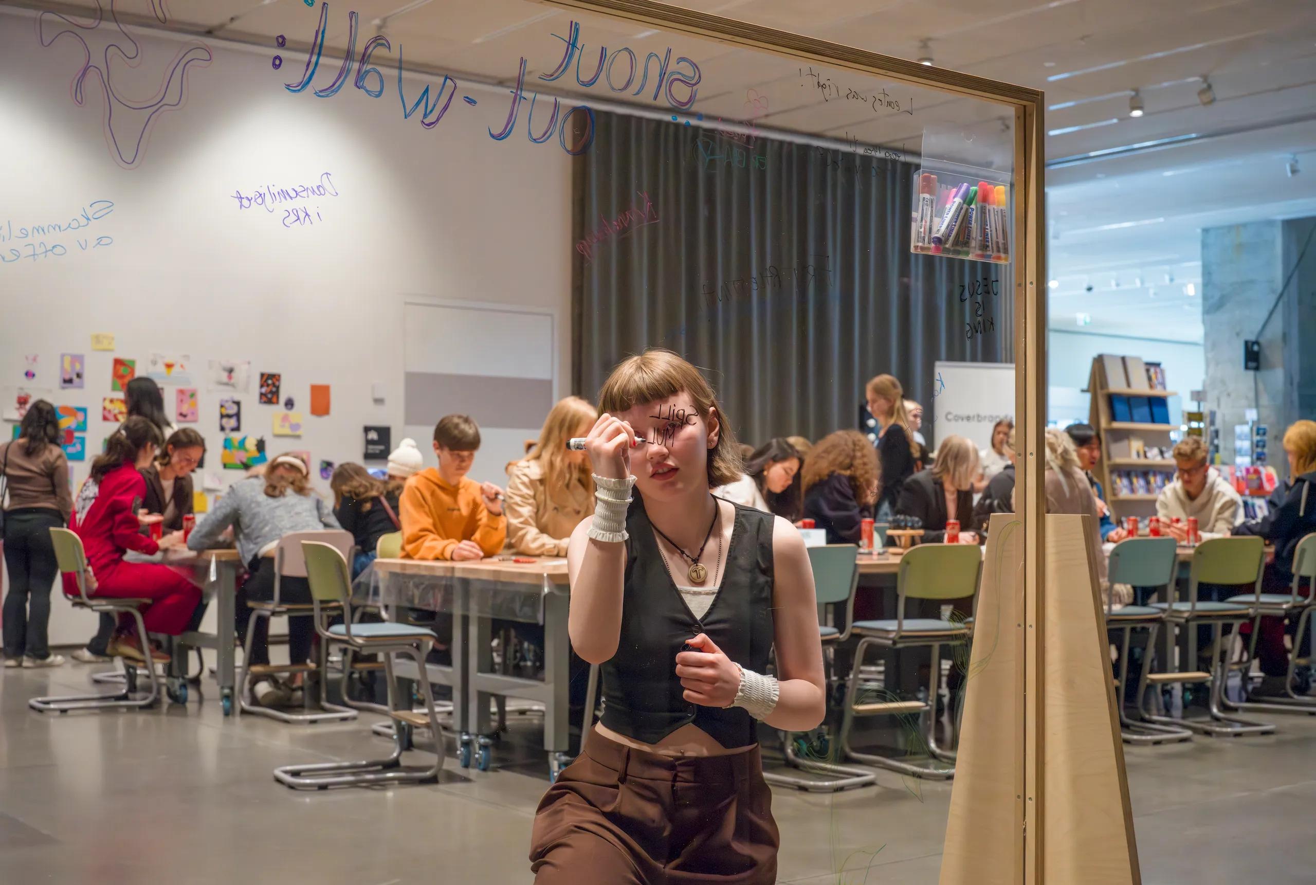 A young woman writes on a transparent board with many people working at tables in the background.