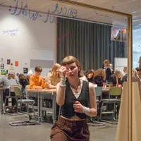 Eine junge Frau schreibt auf eine durchsichtige Tafel, während im Hintergrund viele Menschen an Tischen arbeiten.