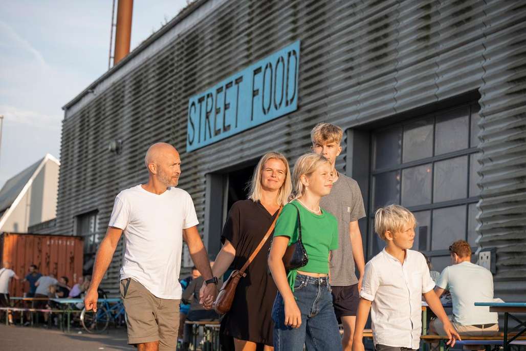 En familie på fem går sammen foran et street food-marked i Aalborg, badet i sollys.