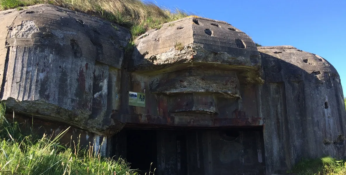 En gammel, delvis nedgravd bunkers fra andre verdenskrig dekket med gress og mose, beliggende i et åpent grønt landskap under en klar blå himmel.