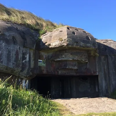 En gammel, delvis nedgravd bunkers fra andre verdenskrig dekket med gress og mose, beliggende i et åpent grønt landskap under en klar blå himmel.