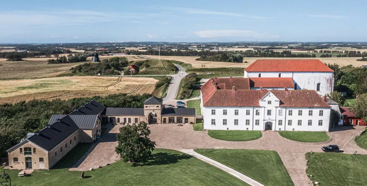 Et vakkert herregårdskompleks sett ovenfra, omgitt av frodige jorder og grønne plener. Hovedbygningen er hvit med rødt tak, og flere mindre bygninger med svarte tak ligger rundt.