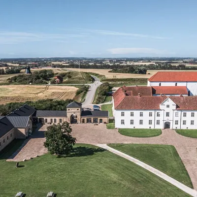 Et vakkert herregårdskompleks sett ovenfra, omgitt av frodige jorder og grønne plener. Hovedbygningen er hvit med rødt tak, og flere mindre bygninger med svarte tak ligger rundt.