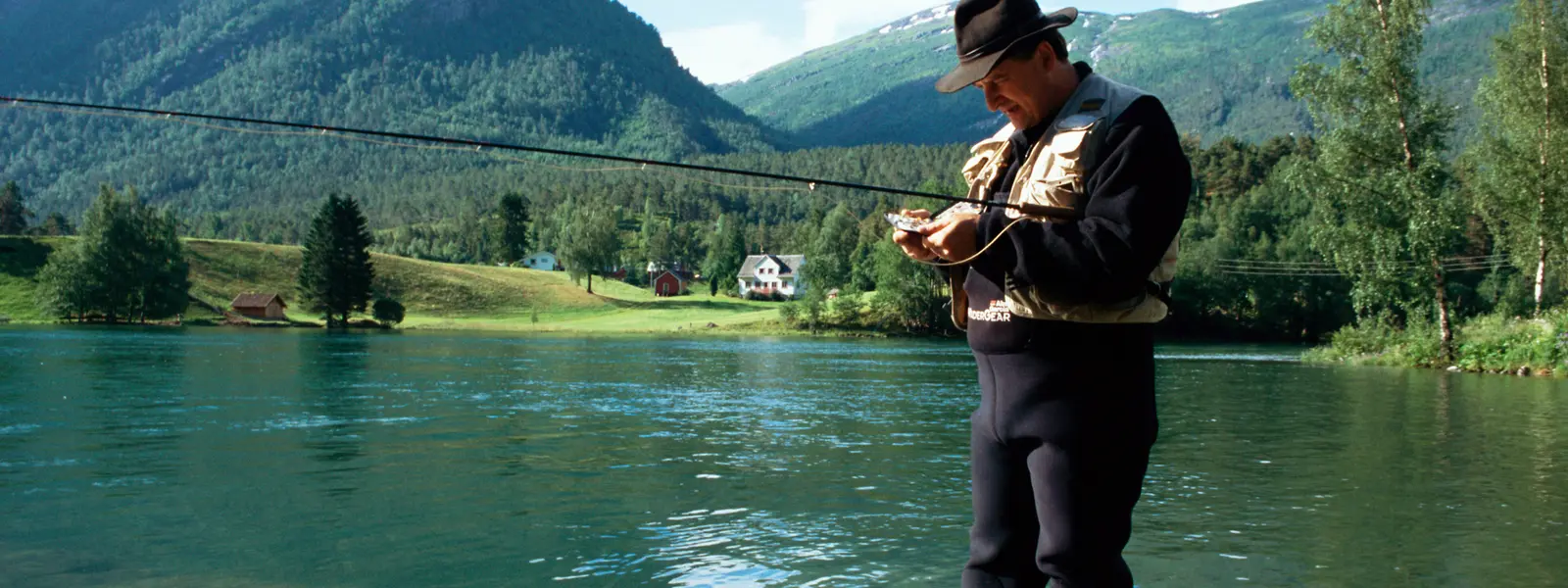 A man fly fishing in a calm river, surrounded by lush green mountains and countryside houses, under a clear sky.