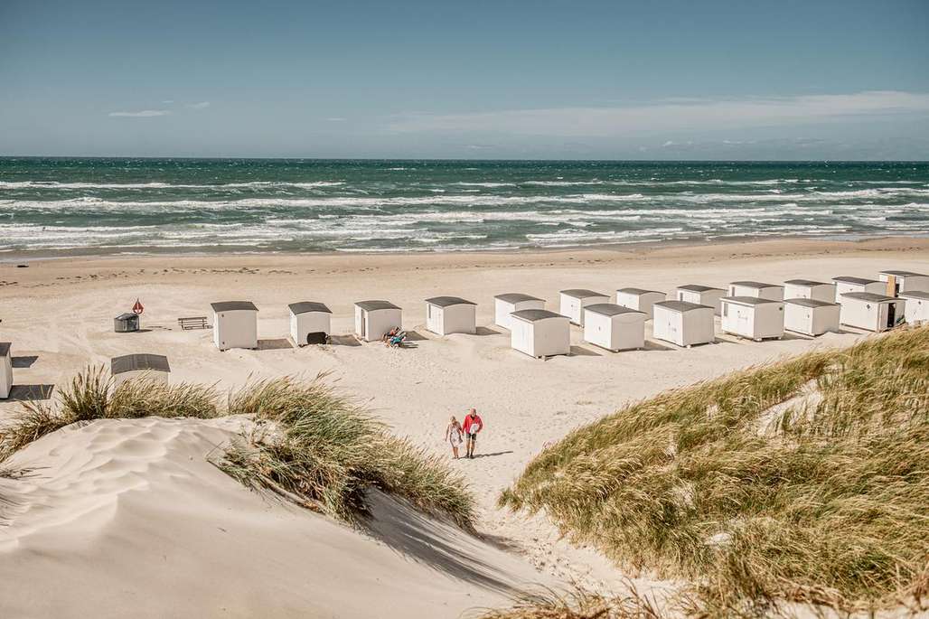 Et par går hånd i hånd langs sanddynene mot stranden. Rader av små, hvite strandhytter står plassert ved vannkanten på en rolig dag ved sjøen.