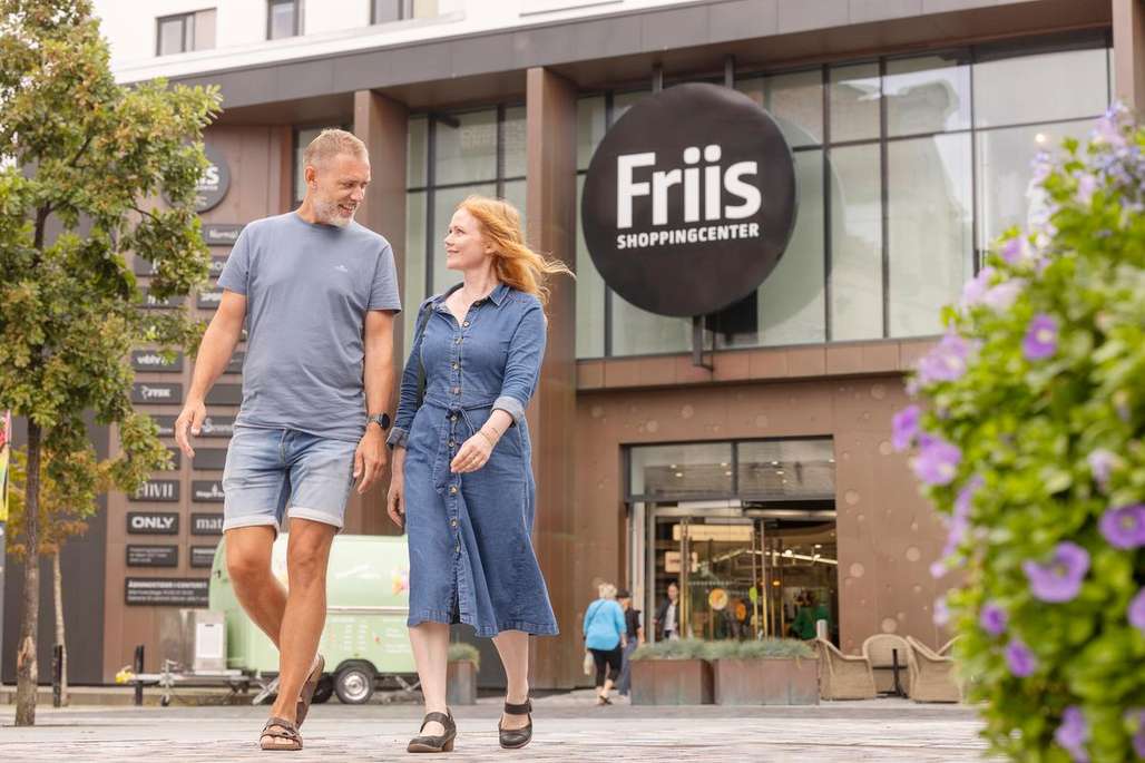 Et smilende par går foran Friis Shoppingcenter i Aalborg, omgitt av grønne trær og blomster.