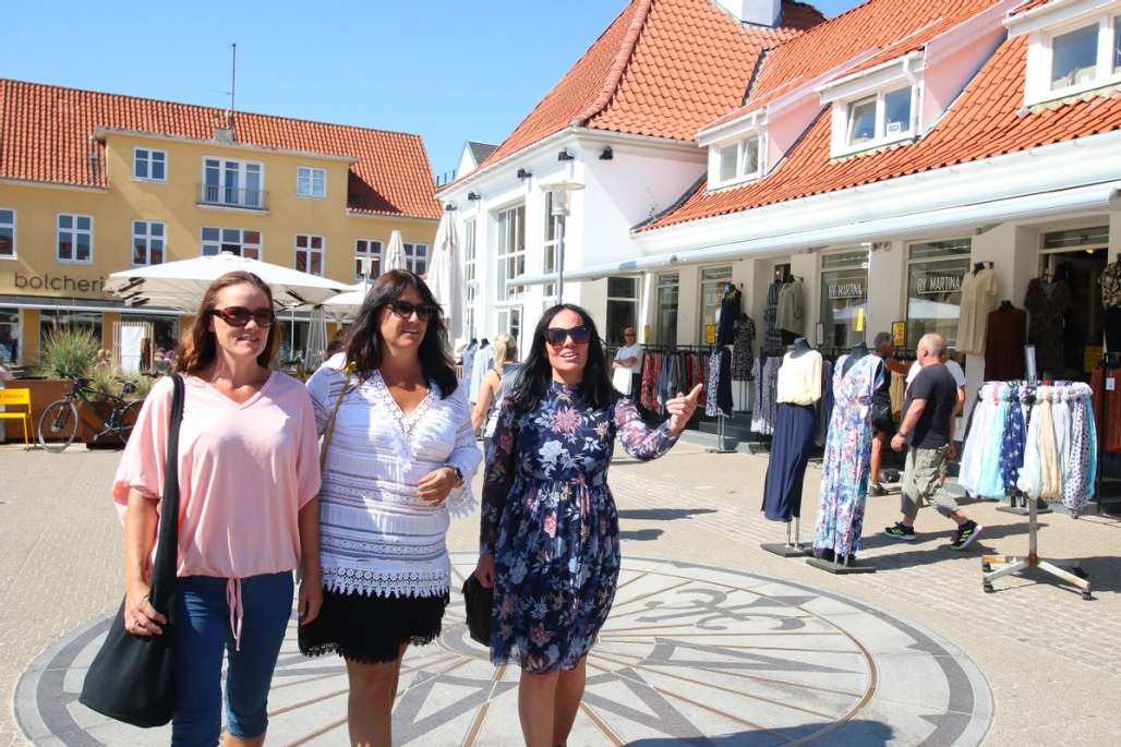 Tre kvinner nyter en solrik dag på shopping i en sjarmerende dansk by med fargerike bygninger og butikker. De går langs en gate fylt med utstillingsdukker og klær.