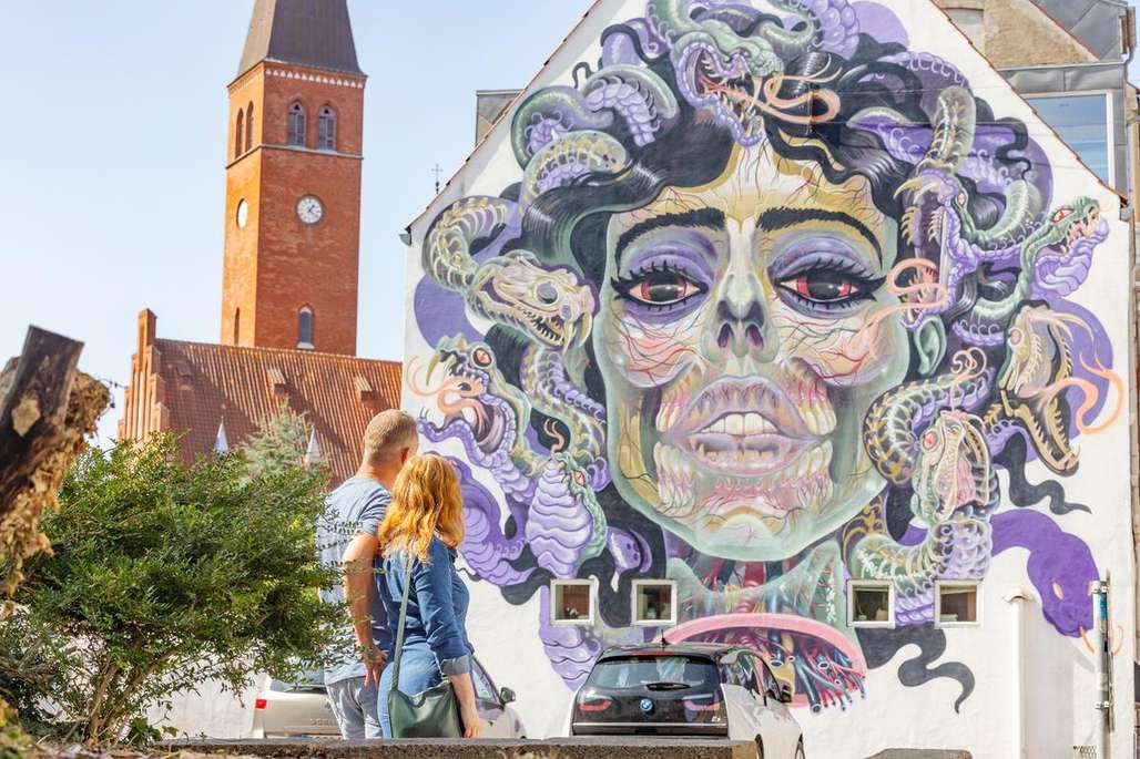 Et par beundrer et fargerikt veggmaleri av en Medusa-figur i Aalborg, med en kirke i bakgrunnen.