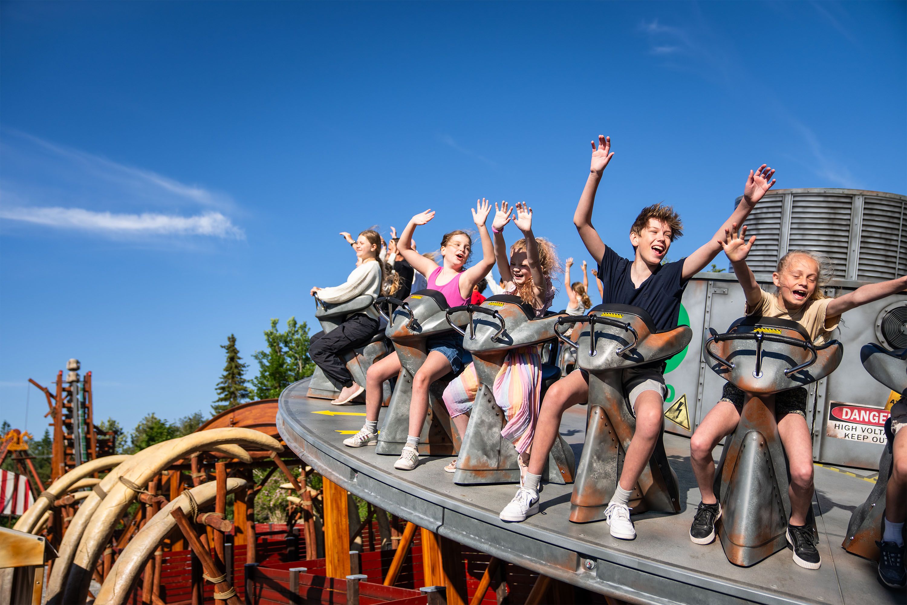 En gruppe barn som sitter på en fornøyelsesparkattraksjon, løfter hendene i været og ler mens de nyter turen, under en klar blå himmel.