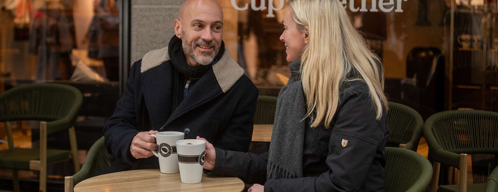 En mann og en dame sitter ute på kafé ved et rundt og drikker kaffi.
