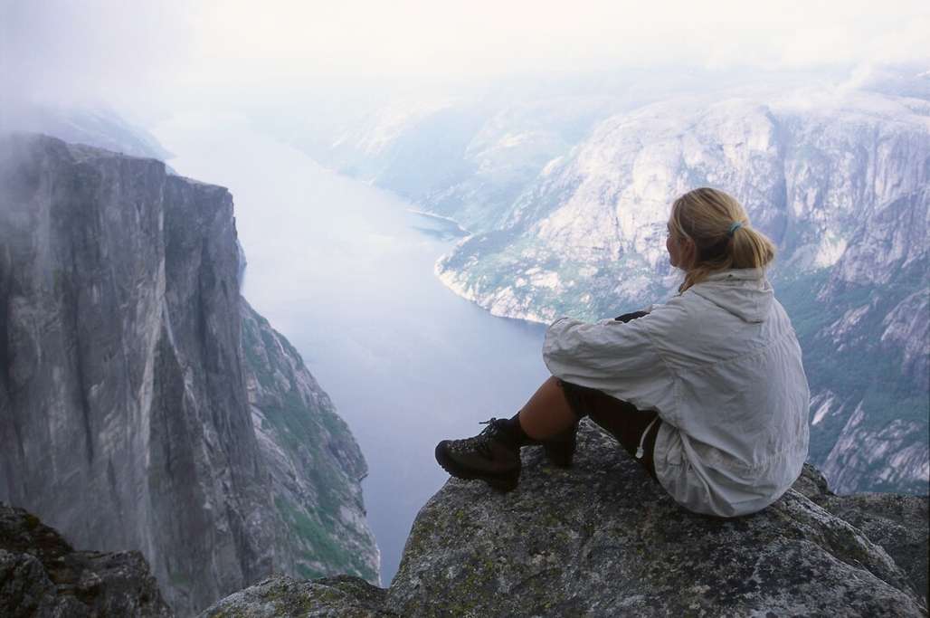 Dame sidder på et bjerg og ser ud over fjord og bjerglandskab.