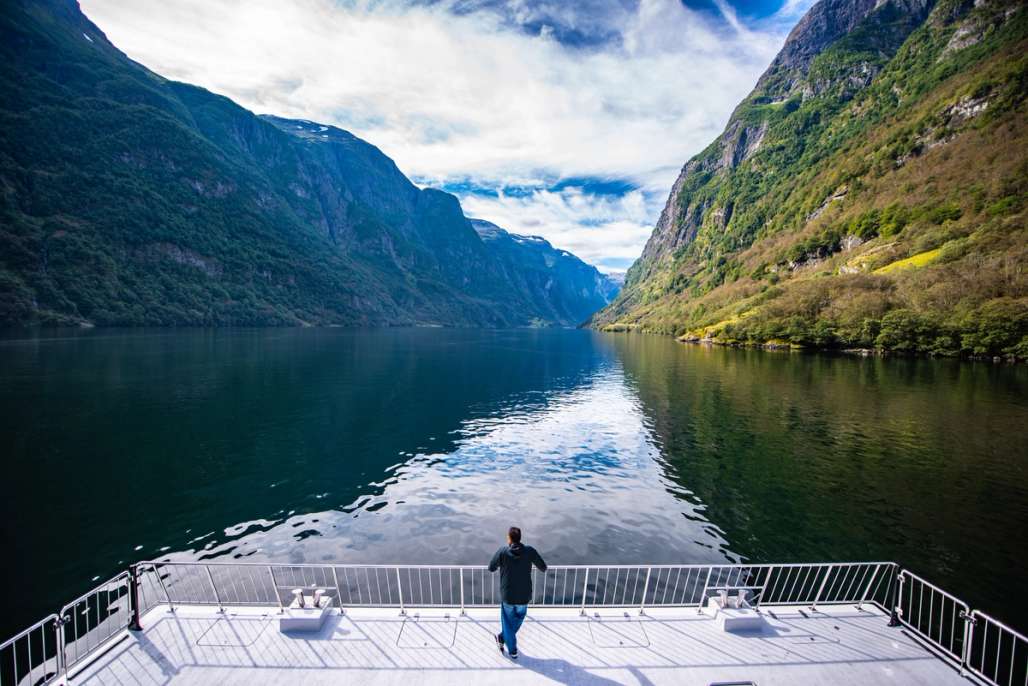 a man is standing on the deck of a boat looking out over a lake surrounded by mountains .