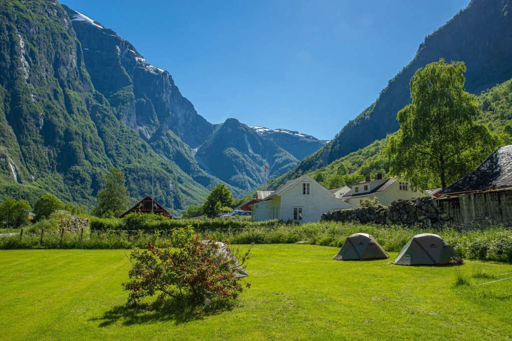 A serene campsite with tents on a lush green lawn, surrounded by towering mountains and traditional houses under a clear blue sky.