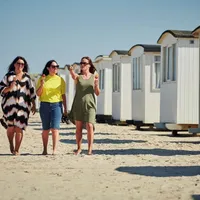 Tre kvinner går langs en sandstrand foran en rekke hvite strandhytter, hver med en is i hånden og smiler mens de prater sammen.