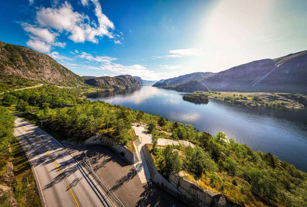 A scenic view of a winding road along a calm fjord, surrounded by steep mountains and lush greenery under a clear blue sky.