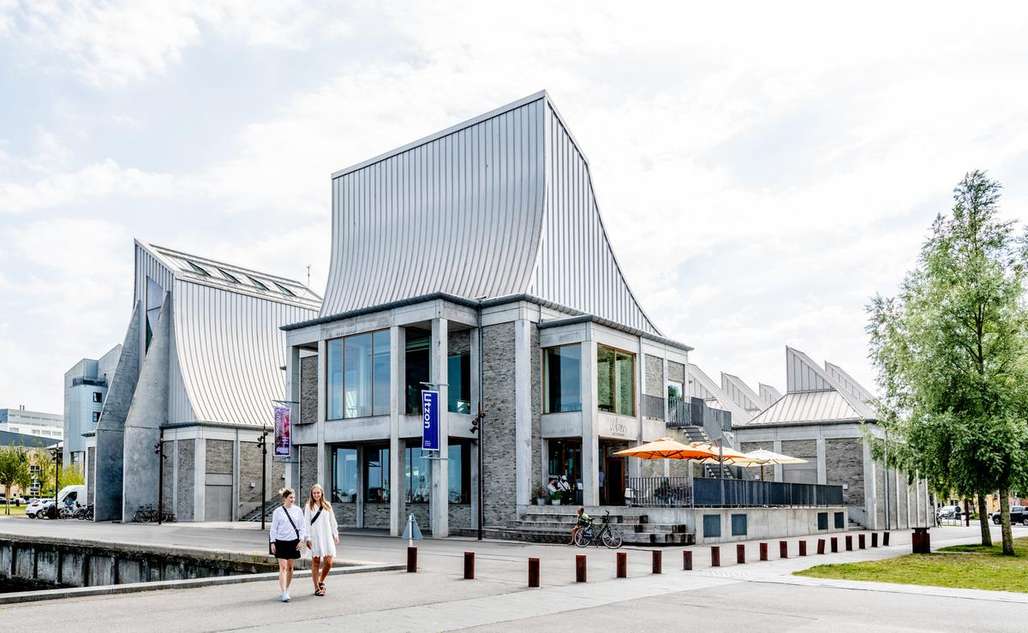 Moderne arkitektur av Utzon Center i Aalborg, med to personer som spaserer utenfor bygningen på en solrik dag.