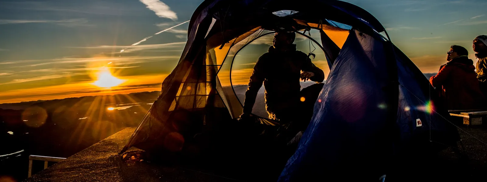 A person sits inside a tent at sunset, with a beautiful view of mountains and valleys in the distance.