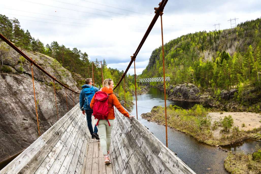 two people are walking across a wooden suspension bridge over a river .