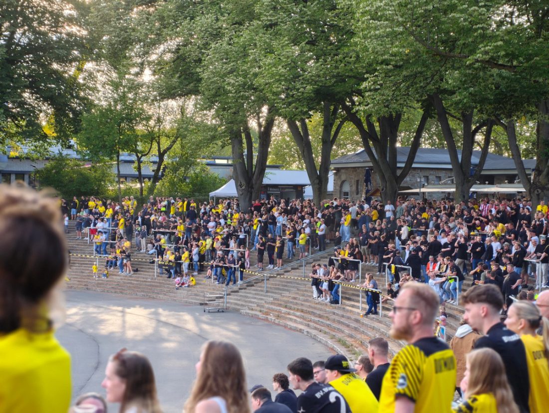 Zuschauer auf den offenen Stehtraversen der Roten Erde zwischen Bäumen und alten Stadiongebäuden.