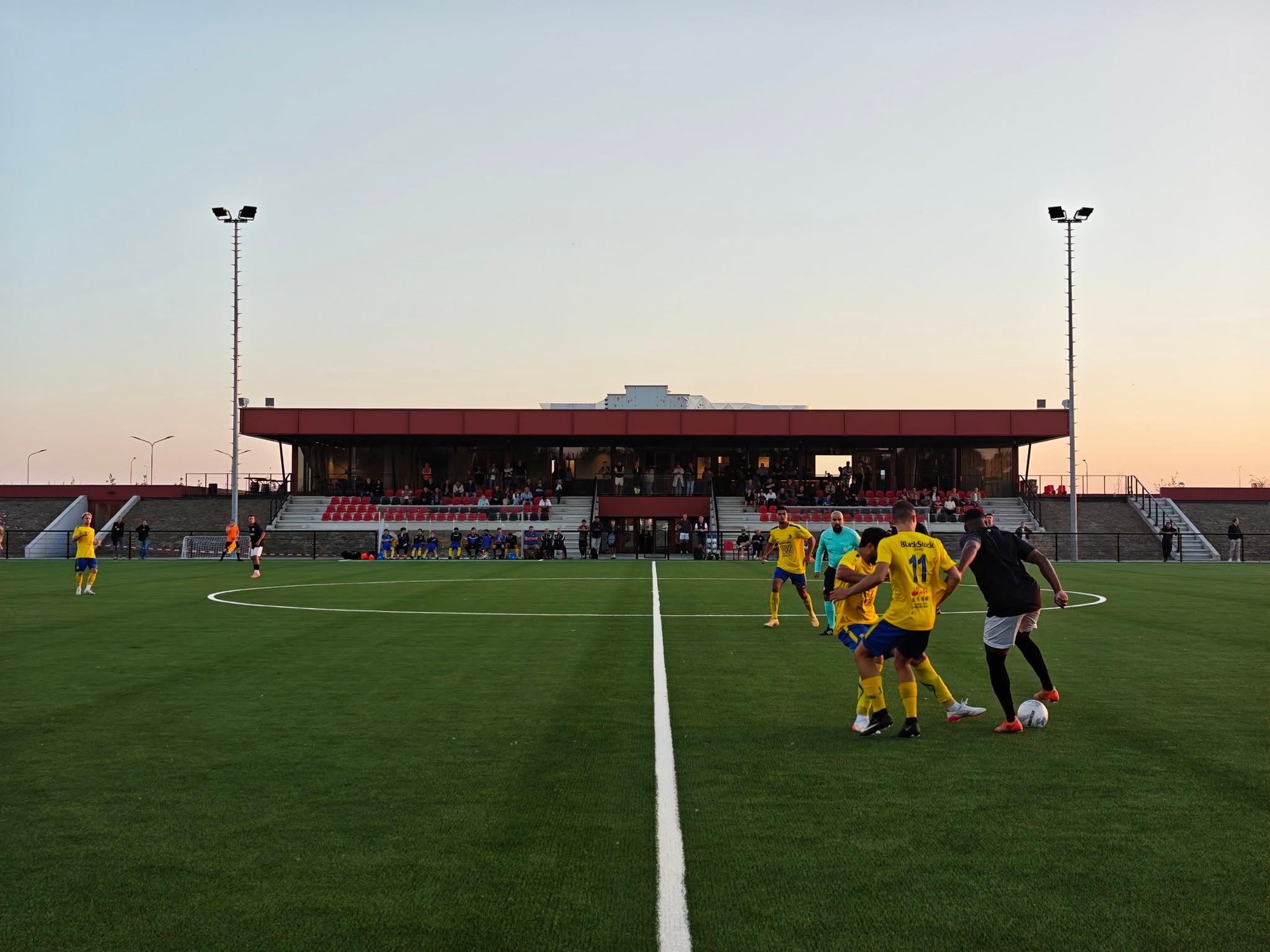 Fußballspiel zwischen SCPB’22 und Legmeervogels auf dem neuen Sportpark De Veldpost in Badhoevedorp. Im Hintergrund die überdachte Haupttribüne im Abendlicht.