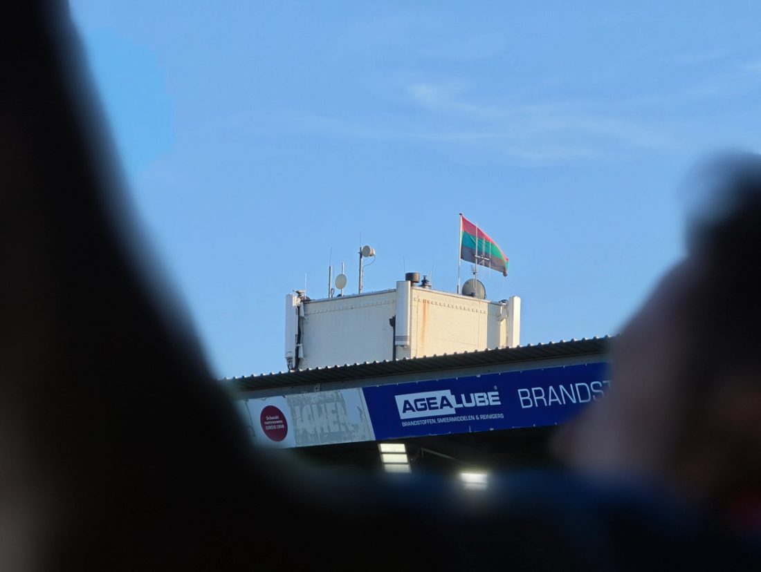Rot-grün-schwarze Fahne auf einem Dach über der Tribüne von De Goffert vor blauem Himmel.
