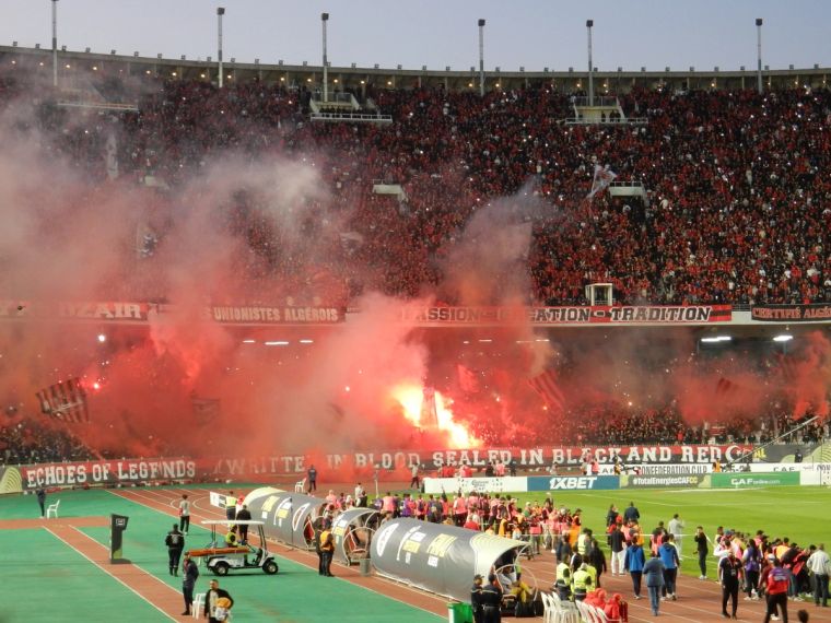 Juillet: volle Ränge und Pyro über der USMA-Kurve. Volles Stade du 5 Juillet bei USM Alger gegen Zamalek, über der roten Fankurve steigen Pyro-Raketen auf.