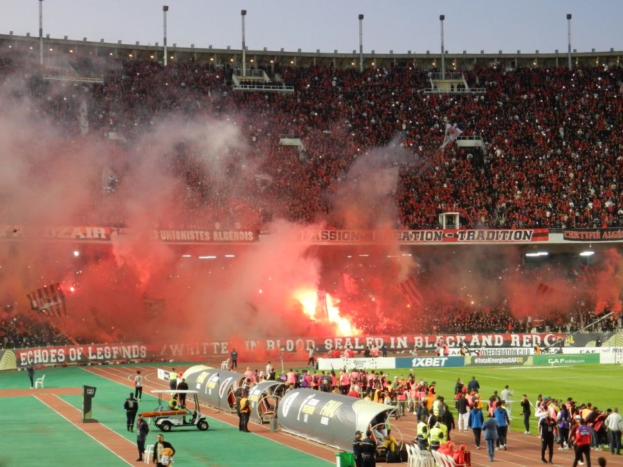 Juillet: volle Ränge und Pyro über der USMA-Kurve. Volles Stade du 5 Juillet bei USM Alger gegen Zamalek, über der roten Fankurve steigen Pyro-Raketen auf.