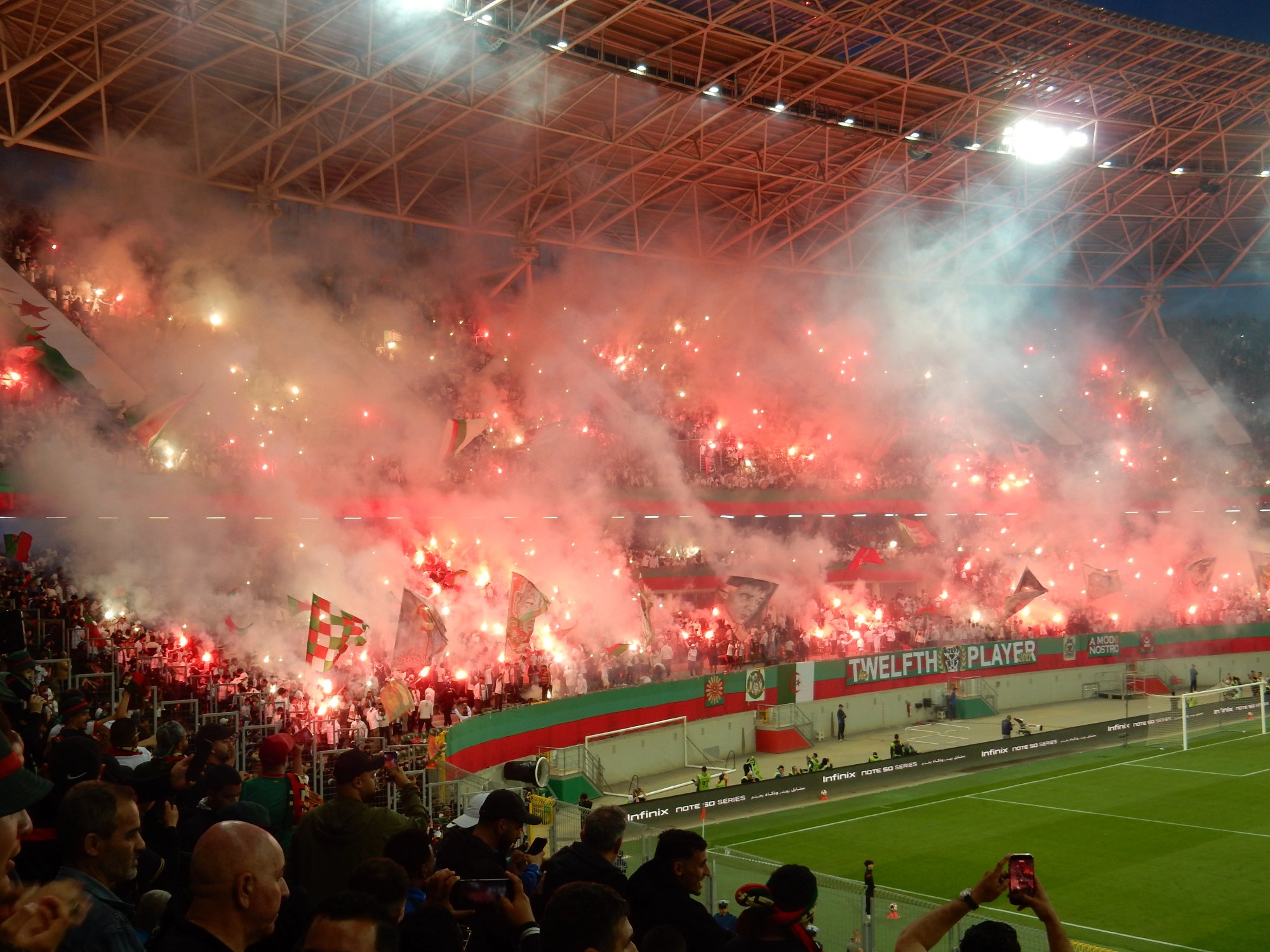 Fans von MC Alger zünden auf mehreren Rängen des Ali-la-Pointe-Stadions zahlreiche rote Fackeln, dichter Rauch zieht unter dem Stadiondach entlang.