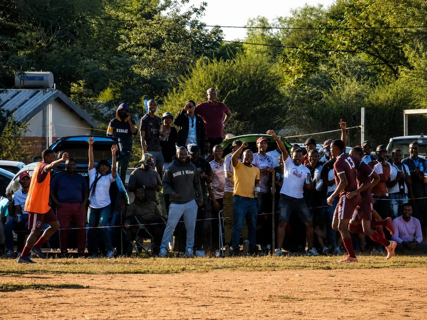 Zuschauer stehen dicht am staubigen Fußballfeld in Mochudi, Botswana, während Spieler der Eastern Tigers in weinroten Trikots an der Seitenlinie vorbeilaufen.