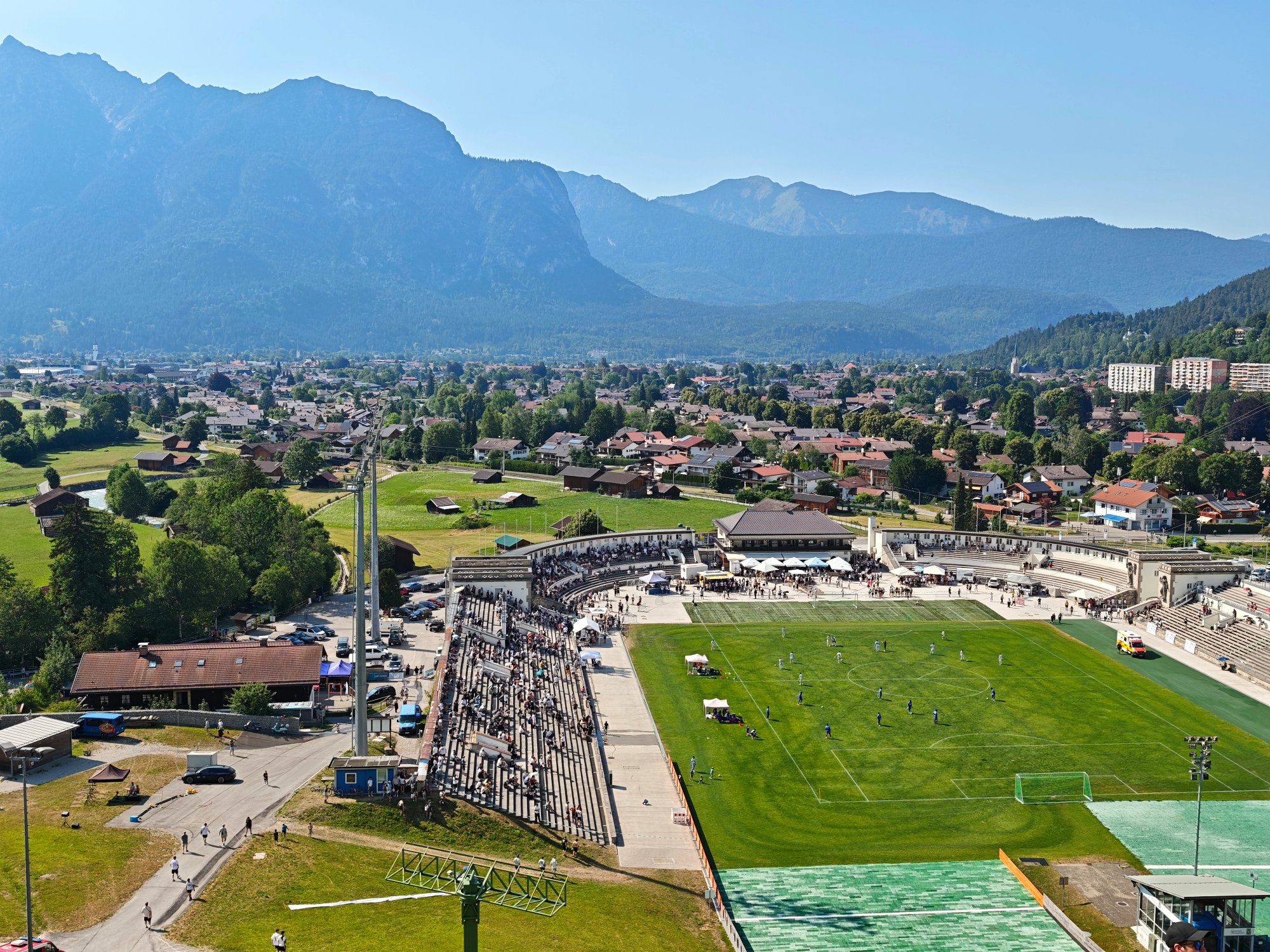 Blick von oben auf das Olympiastadion in Garmisch-Partenkirchen mit Fußballspiel, Tribünen und Bergpanorama im Hintergrund.